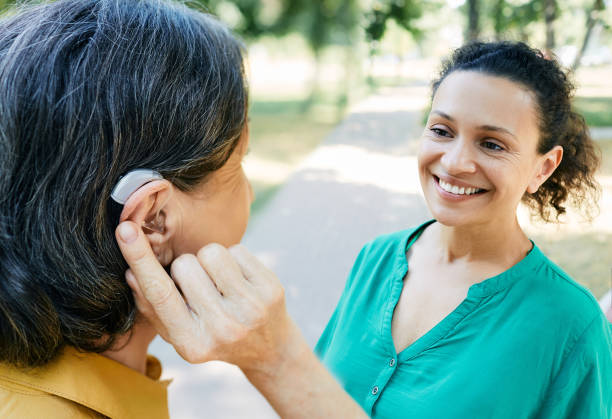 Зрелая женщина с нарушением слуха использует слуховой аппарат для общения со своей подругой на открытом воздухе. Слуховые решения
